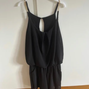 Svart kort klänning med inbyggda shorts med guldkedjor - Snygg svart kort klänning  med inbyggda shorts, tunna axelband dekorerade med guldfärgade kedjor. Klänningen har en lös passform upptill, markerad midja och en liten öppning i ryggen. Perfekt för dig som gillar stilrena detaljer och en chic look.