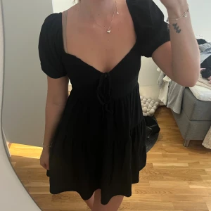 Svart kort klänning med puffärm - Supersöt svart kort klänning med puffärmar och v-ringning. Klänningen har knyt framtill och en volangig kjol som ger en flowy känsla. Perfekt för dig som gillar en feminin och trendig stil. Materialet känns lätt och bekvämt mot huden.