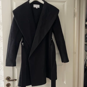 Svart kappa från Vila med bälte - Stilren svart kappa från Vila med bred krage och knytbälte i midjan. Långärmad modell i mjukt ullmaterial, perfekt för kyliga dagar. Snygg och enkel design som ger en clean look.