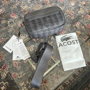 Grå axelväska från Lacoste - Snygg grå axelväska från Lacoste med geometriskt mönster och silverfärgad logga framtill. Väskan har justerbar axelrem, dragkedja och detaljer i syntetmaterial. Perfekt storlek för det viktigaste och stilren design med diskreta Lacoste-detaljer.