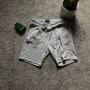 Grå mjuka shorts från Lyle & Scott - Säljer ett par grå shorts från Lyle & Scott Junior med elastisk midja och fickor fram och bak. Materialet är mjuk bomull och på ena benet finns den klassiska gula fågel-loggan broderad. Perfekta för chill dagar och sommarhäng.