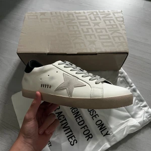 Golden Goose vita sneakers med stjärna - Golden Goose sneakers i vitt skinn med grå mockadetaljer och ikonisk stjärna på sidan. Skorna har grå snörning, svart hälparti och beige sula. Klassisk låg modell med rund tå och platt sula, perfekt för en clean och trendig look.