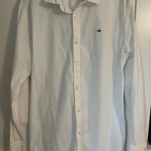 Vit skjorta från Tommy Jeans (liten i storlek) - Klassisk vit skjorta från Tommy Jeans i slim fit-modell. Skjortan har långa ärmar, knäppning framtill och en liten broderad logga på bröstet. Tillverkad i bomull för en fräsch och stilren look. Liten i storlek.