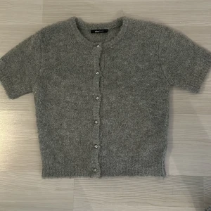 Grå stickad kofta från Gina Tricot - Mysig grå stickad kofta från Gina Tricot i storlek XS. Koftan har korta ärmar, rund hals och silverfärgade knappar framtill. Materialet är fluffigt och ger en skön känsla. Perfekt för dig som gillar enkel och stilren design.