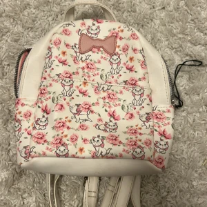 Söt ryggsäck från Disney  - Supergullig liten ryggsäck från Disney med vita och rosa nyanser, täckt av blommor och katter samt en rosa rosett framtill. Väskan har dragkedja och är fodrad med rosa tyg. Perfekt för dig som gillar söta detaljer och vill sticka ut. (Priset kan diskuteras)