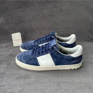 Valentino Garavani blå sneakers mocka - Snygga sneakers från Valentino Garavani i blå mocka med vita skinnpaneler och vita detaljer vid häl och sula. Skorna har snörning och coola nitar baktill på sulan för en extra edge. Perfekta för dig som vill ha något unikt och stilrent.