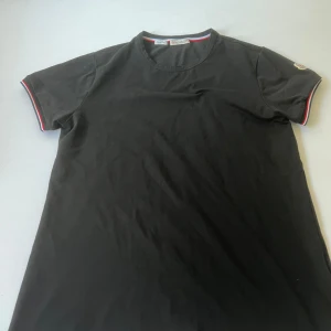 Svart Moncler t-shirt med logga - Svart t-shirt från Moncler i bomull med korta ärmar. Ärmsluten har snygga ränder i rött, vitt och blått samt en broderad Moncler-logga på ena ärmen. Klassisk rund halsringning och normal passform. Perfekt för en clean och stilren look.
