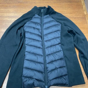 Svart hybridjacka med dragkedja - Snygg svart jacka med quiltad, vadderad framsida och släta ärmar. Jackan har hel dragkedja framtill och en sportig look som passar perfekt till lager-på-lager. Enkel och stilren design med hög krage.