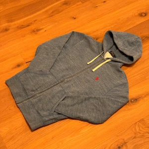 Ljusblå hoodie från Polo Ralph Lauren - Mysig ljusblå (ursäkta vilseledande bilder) hoodie från Polo Ralph Lauren med dragkedja, huva och vita snören. Tröjan har en röd broderad logga på bröstet och praktiska fickor framtill. 