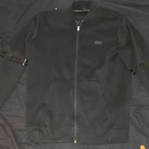 Svart ziptröja från BOSS i medium - Svart långärmad ziptröja från BOSS med diskret logga på bröstet och svarta glansiga detaljer på ärmarna. Tröjan har ribbade muddar, ståkrage och två fickor framtill. Perfekt för en clean och stilren look. 2 bokstäver saknas