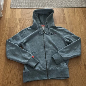 Blå hoodie från Superdry Orange Label - Säljer en grå hoodie från Superdry Orange Label med dragkedja, broderad logga på bröstet och huva med snörning. Tröjan har två fickor framtill och ribbade muddar. Perfekt för chill dagar och streetstyle.