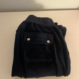 Mörkblå velourbyxor Juicy Couture S - Mörkblå mjukisbyxor från Juicy Couture i velour med bred resår i midjan och två bakfickor med lock och guldfärgade knappar. Klassisk 00-talsstil, perfekta för en chill och trendig look.