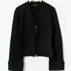 Svart kofta från Gina Tricot - Figursydd kofta med intagen midja. Koftan är svart och har uppvikta ärmar med knappar. Den har v-ringning, lång ärm, knappar framtill och stickat material. Bara testad och är helt oanvänd, men utan prislapp.