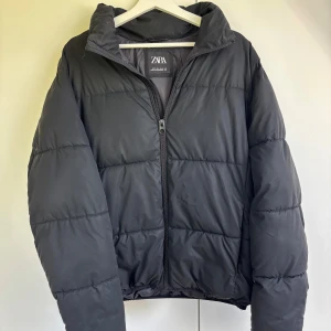Svart pufferjacka från Zara, M - Svart pufferjacka från Zara i storlek M. Jackan har hög krage, dragkedja framtill och quiltad design. Tillverkad i syntetmaterial och perfekt för kyliga dagar. Enkel och stilren look som passar till det mesta.
