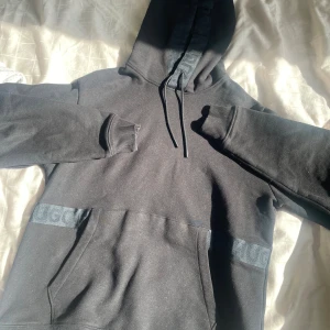 Svart hoodie från HUGO med logga - Svart hoodie från HUGO med broderad logga på huvan och ett band med HUGO-tryck över magen. Tröjan har känguruficka, snörning i huvan och ribbade muddar. Perfekt för en chill och stilren look. Köpt för 799:- 