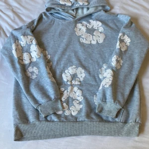 Grå hoodie från Denim Tears - Säljer en grå hoodie från Denim Tears med vita molnliknande tryck över hela tröjan. Hoodien har huva, magficka och ribbade muddar. Tillverkad i mjuk bomull och perfekt för dig som gillar streetwear med unik design.