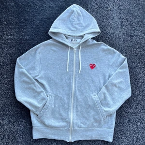 CDG Zip Hoodie  - Grå hoodie med dragkedja från Comme des Garçons Play. Klassisk modell med huva, vita snören och det ikoniska röda hjärtat med ögon broderat på bröstet. Mjuk bomullskvalitet och kängurufickor framtill. Perfekt för en chill och stilren look. Skick 9/10, storlek M men passar även S, äkta. Pris går att diskutera✅