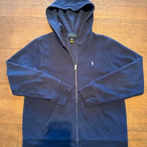 Marinblå hoodie från Polo Ralph Lauren - Snygg marinblå hoodie från Polo Ralph Lauren med dragkedja och huva. Framtill finns en liten broderad logga och på ryggen ett stort tryck med POLO 67. Praktiska fickor och mjukt bomullsmaterial gör den perfekt för chill dagar. Storlek 14-16 år. Nypris ligger runt 1200 och den är i bra skick. Hör av er vid frågor. 