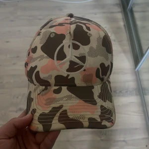 Mercedes x Monster camo keps - Säljer en keps med camouflagemönster i beige, brun och rosa toner. Kepsen har Mercedes-logga broderad fram och bak samt Monster Energy-logga på insidan. Justerbar rem bak för perfekt passform. Perfekt accessoar för dig som gillar streetstyle och motorsport.