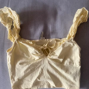 Gul linnetopp med spets och knyt - Supersöt ljusgul linnetopp från NA-KD med breda axelband i spets och en liten knytning framtill. Toppen är croppad och har en romantisk vibe med fina spetsdetaljer längs kanterna. Perfekt för varma dagar eller lager på lager.