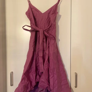 Rosa omlottklänning från Vila - Supersöt rosa klänning från Vila med smala axelband, v-ringning och omlottknytning i midjan. Klänningen har en flowy, asymmetrisk kjol och smockad rygg för extra komfort. Perfekt för dig som gillar en feminin och trendig stil.