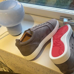 Gråa Christian Louboutin sneakers - Snygga grå sneakers från Christian Louboutin i mocka med klassisk röd sula och vita kanter. Skorna har rund tå, snörning och en stilren design som passar perfekt till streetwear. Insidan är fodrad i beige och sulan har Louboutins ikoniska signatur.