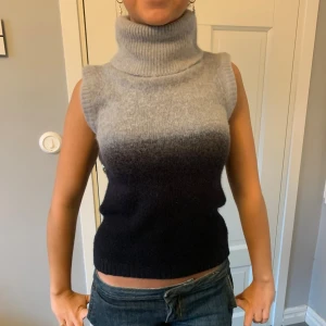 Ärmlös polotopp i grått och svart - Stickad ärmlös polotopp med hög krage och snygg ombre-effekt från ljusgrått upptill till svart nedtill. Mjuk och skön, perfekt att styla med jeans eller kjol. Ribbstickade kanter vid ärmhål och hals. 