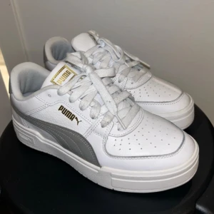Vita och grå sneakers från Puma - Snygga vita sneakers från Puma med grå detaljer och guldfärgade loggor. Skorna har klassisk låg modell, snörning och är tillverkade i skinn och syntet. Perfekta för dig som gillar stilrena och fräscha sneakers med en sportig vibe. Skorna är i topp skick då de är knappt använda. Skorna är i storlek 40.5 och anses vara normala i storleken.