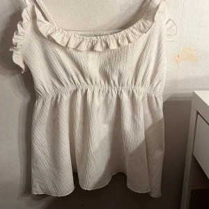 Vit volangtop från 157, XS - Supersöt vit linnetopp från 157 i storlek XS. Toppen har smala axelband med knyt, volangdetaljer vid ringningen och är tillverkad i ett lätt, krinklat tyg. Perfekt för varma dagar och ger en luftig känsla med sin avslappnade passform. Använd 1 gång!