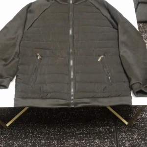 Svart quiltad pufferjacka med dragkedja och luva - Svart pufferjacka med quiltad framsida och två snedställda fickor med dragkedja. Jackan har hel dragkedja framtill och hög krage. Materialet är troligtvis polyester och jackan är långärmad med en clean, stilren look. Kan anvädas inomhus och ute!