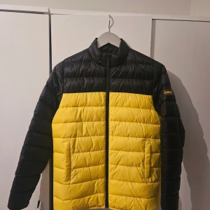 Svart och gul pufferjacka Barbour XL - Säljer en fet pufferjacka från Barbour International i svart och gul. Jackan har hög krage, två sidofickor och dragkedja framtill med Barbour-logga. Perfekt för dig som vill sticka ut med en cool färgblockad look. Jackan är vadderad och riktigt skön när det är kallt ute och använd 2 gånger (storlek XL Junior)