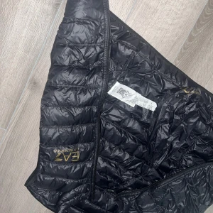Svart dunjacka från EA7 Emporio Armani - Säljer en svart dunjacka från EA7 Emporio Armani med guldfärgad logga på bröstet och insidan. Jackan har quiltad design, dragkedja framtill och lätt glansigt material. Perfekt för dig som vill ha en snygg och stilren jacka med exklusiv känsla.