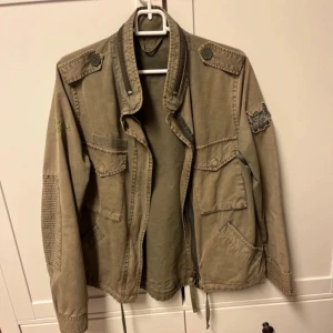 Grön Odd Molly jacka  - Säljer en grön Odd Molly jacka med snygga knappar på axlarna, broderad patch på ärmen och flera praktiska fickor. Jackan har dragkedja framtill, snörning i nederkant och ribbade detaljer på ärmarna. Perfekt för inför hösten men även våren. Fint skick.