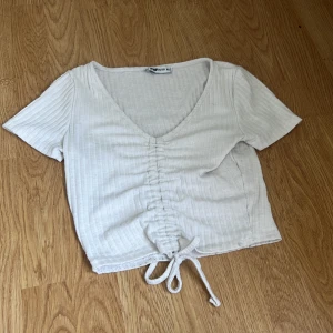 Vit ribbad croppad topp med snörning - Säljer en vit croppad topp från Bik Bok med ribbad struktur och snygg snörning framtill. Toppen har korta ärmar, v-ringad hals och en omlott-effekt vid midjan. Perfekt för dig som gillar stilrena och trendiga plagg.