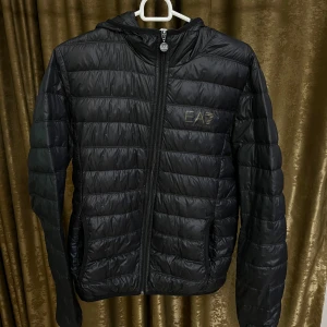 Dunjacka från EA7 Emporio Armani - Säljer en svart dunjacka från EA7 Emporio Armani med huva och dragkedja. Jackan har quiltad design och logga i guld på bröstet. Perfekt för kyliga dagar och riktigt snygg till streetwear-stilen.