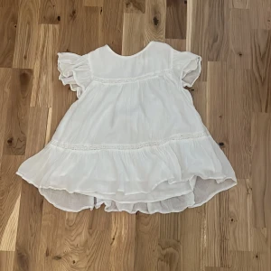 Vit volangklänning från Gina Tricot - Superfin vit blus från Gina Tricot med volanger och korta, luftiga ärmar. Blusen har en flowy passform och är tillverkad i ett lätt tyg som känns perfekt för varmare dagar. Stängs med knapp i nacken. 