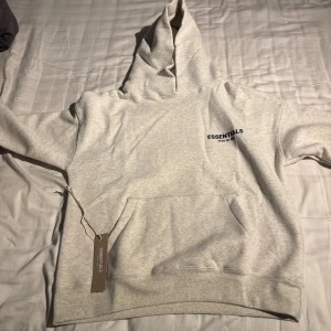 Ljusgrå Essentials Fear of God hoodie - Ljusgrå hoodie från Essentials Fear of God med svart logga på bröstet och ryggen. Klassisk känguruficka framtill, stor huva och ribbade muddar. Mjuk och skön bomullsblandning, perfekt för chill dagar. Snygg streetwear-vibe och enkel att matcha.