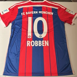 Bayern München Robben 10 matchtröja - Säljer en officiell FC Bayern München fotbollströja från säsongen 2014/2015 med Robben 10 på ryggen. Tröjan är Adidas ClimaCool, har röda och blå vertikala ränder, vita detaljer och Bundesliga-märke på ärmen. Perfekt för dig som älskar tysk fotboll och klassiska matchtröjor. Replika, endast handtvätt gäller!