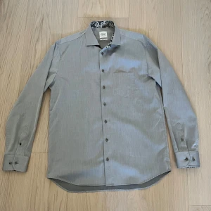 Grå skjorta från Oscar of Sweden - Snygg grå skjorta från Oscar of Sweden i regular fit. Skjortan har lång ärm, bröstficka och coolt blommigt mönster på insidan av kragen. Tillverkad i bomull och är strykfri. Perfekt för dig som gillar stilrena plagg med en twist. Storlek 42 och aldrig använd.