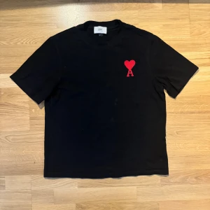 Svart t-shirt från Ami Paris med rött hjärta - Svart t-shirt från Ami Paris med klassisk passform och korta ärmar. På bröstet finns ett broderat rött hjärta med bokstaven A. Tillverkad i mjuk bomull, perfekt för en clean och stilren look.