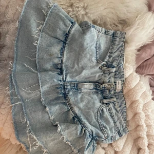 Ljusblå volangkjol i jeans från Perfect Jeans - Supersöt ljusblå jeanskjol från Perfect Jeans med volanger och råa, fransiga kanter. Kjolen har klassiska fickor fram och bak, bälteshällor och dragkedja med knapp. Perfekt för dig som vill ha en trendig och avslappnad look.