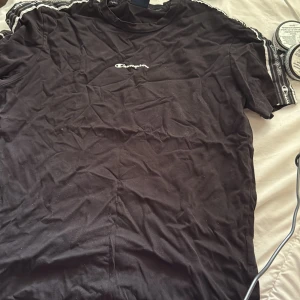 Svart Champion t-shirt med logga - Svart t-shirt från Champion i bomull med klassisk logga broderad på bröstet och loggoband längs axlarna. T-shirten har rund hals och korta ärmar, perfekt för en sportig och avslappnad stil.