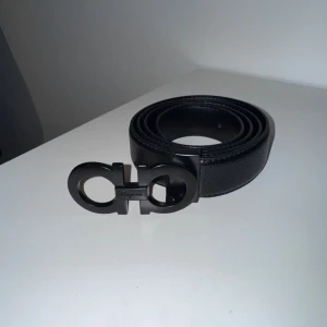 Svart Ferragamo skärp - Säljer ett svart skärp från Ferragamo i skinn med en ikonisk dubbel Gancini-spänne i matt svart metall. Bältet har en stilren och klassisk design som passar till jeans eller kostymbyxor. Perfekt accessoar för att lyfta din outfit. (110cm)