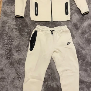 Beige Nike hoodie med dragkedja XL - Nike hoodie i beige med svart logga och svarta detaljer vid fickorna. Tröjan har huva, hel dragkedja och långa ärmar. Materialet är mjukt och känns som bomull eller polyester, perfekt för chill eller träning. Snygg kontrast med svarta detaljer och stilren design.