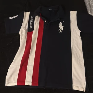 Randig Polo Ralph Lauren t-shirt L - Snygg t-shirt från Polo Ralph Lauren i storlek L. Modellen har korta ärmar, klassisk krage och knappar vid halsen. Färgerna är marinblå, röd och vit med breda ränder framtill och stor broderad logga på bröstet. Tillverkad i mjuk bomull för skön känsla.