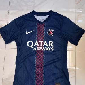PSG Tröja Player Verision  - Paris Saint-Germain fotbollströja med O. Dembélé och nummer 10 på ryggen. Djupblå färg med röd och vit rand längs mitten, PSG-emblem på bröstet och Qatar Airways-tryck. Tillverkad i lätt Dri-FIT-material, perfekt för fotboll eller supporterliv.
