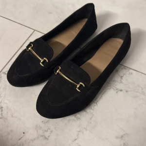 Svarta mockaloafers med gulddetalj - Stilrena svarta loafers i mocka med snygg guldfärgad metallspänne över foten. Skorna har en klassisk, rund tå och platt sula, vilket gör dem både bekväma och trendiga. Använda men fortfarande fräscha! Köparen står för frakten & paketet skickas per post :)