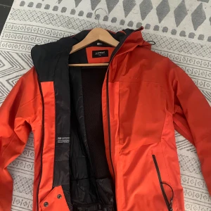 Röd skidjacka från Everest - Snygg röd skidjacka från Everest med svart innerfoder och meshpanel. Jackan har huva, dragkedja framtill och snedställd ficka med dragkedja. Perfekt för skidagar och outdoor-aktiviteter. Sportig look och praktisk design.