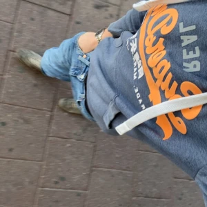 Superdry hoodie  - Säljer en sjukt fet och unik hoodie från Superdry med orange och vitt tryck på bröstet. Tröjan har huva med vita snören och en klassisk känguruficka framtill. Perfekt för dig som gillar streetwear och vill ha en snygg och bekväm look.
