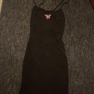 Svart kortklänning med fjäril - Svart figurnära kortklänning med tunna axelband och en rosa fjäril på bröstet. Klänningen har snörning i ryggen för en snygg passform och är perfekt för dig som gillar stilrena detaljer med en twist. Materialet känns mjukt och stretchigt.